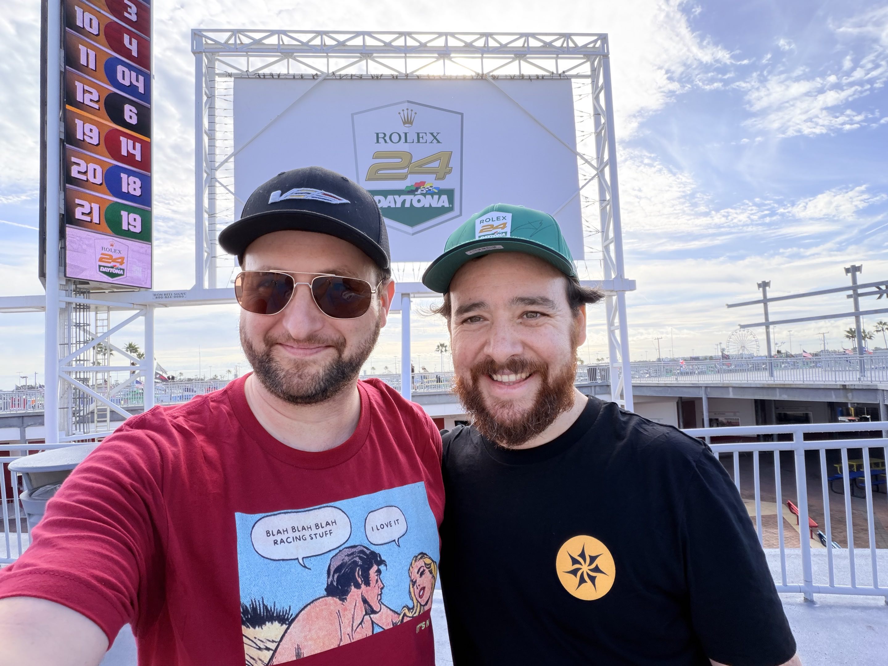 Two men in racing gear smiling for a selfie in front of the Daytona International Speedway timing tower on the garage roof